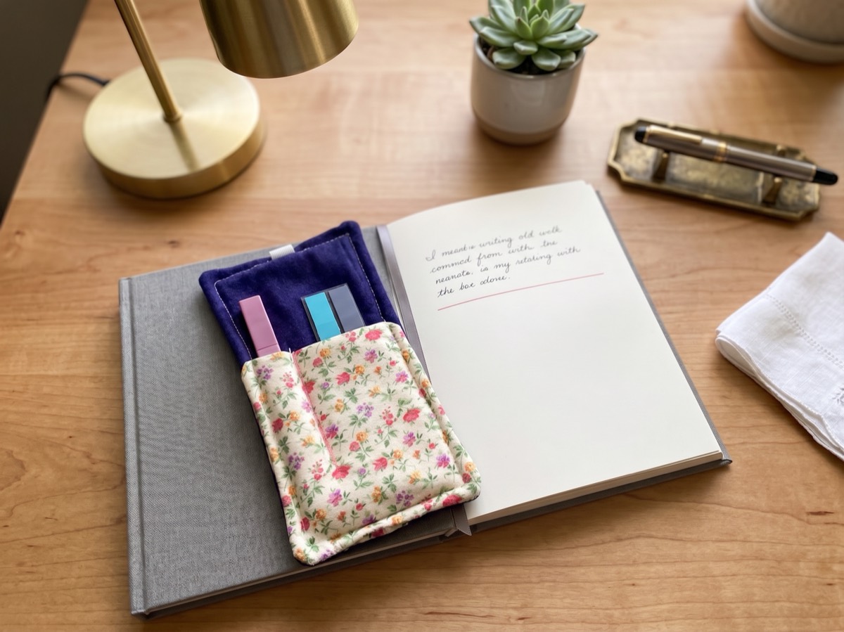 A small floral fabric pen pocket holding three pens, strapped to an open notebook on a wooden desk beside a brass lamp.