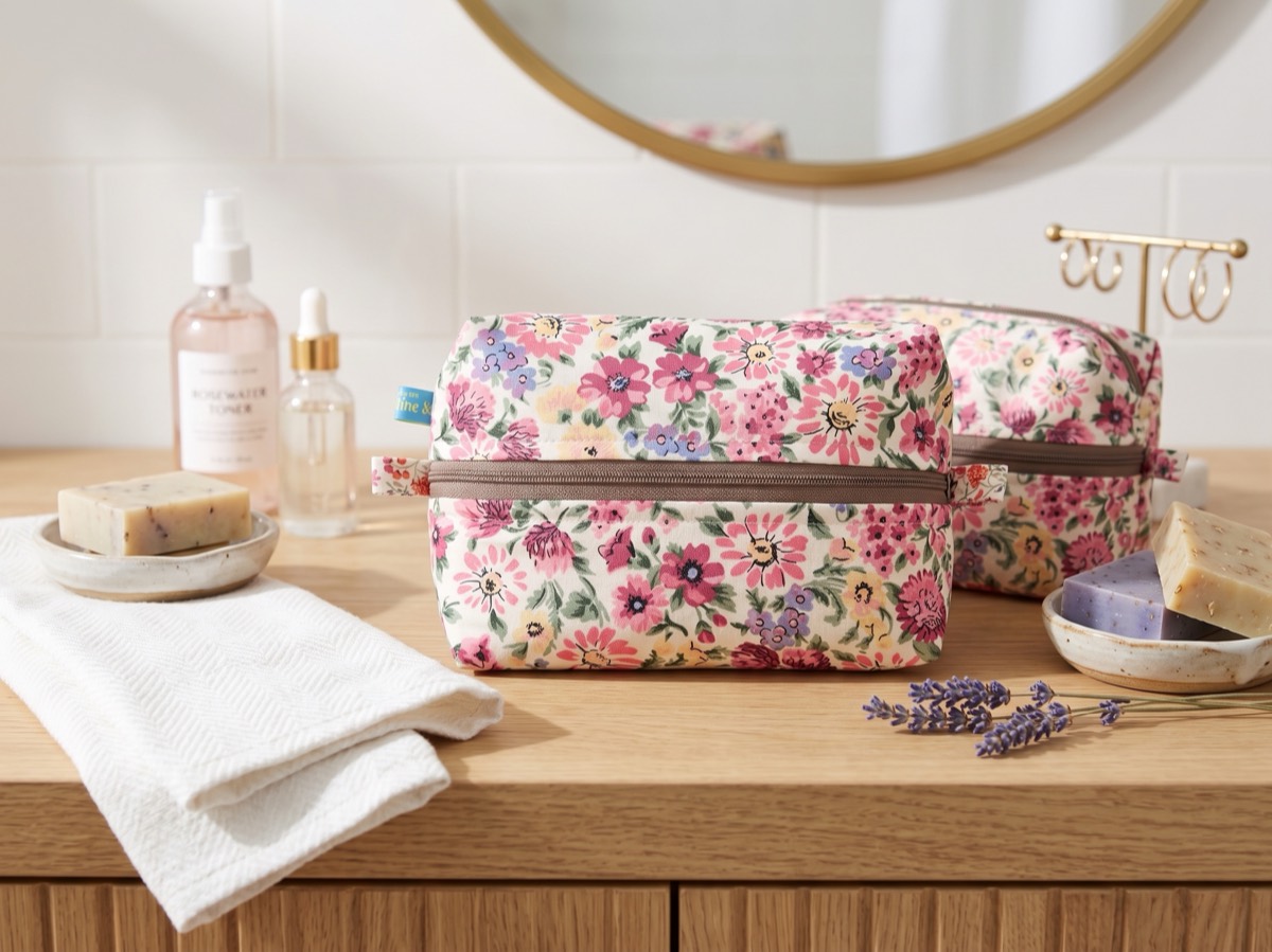A floral zip-top utility bag on a wooden bathroom counter, beside bars of soap, a rosewater bottle and sprigs of lavender.